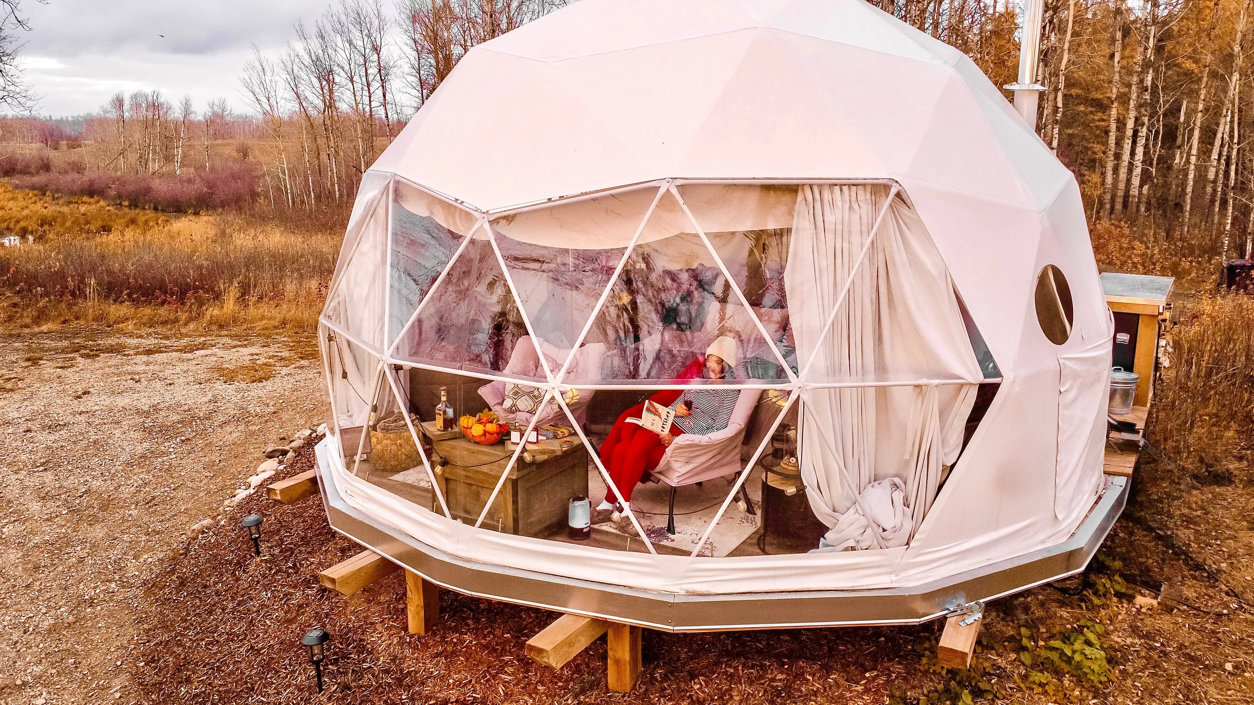 exterior shot of ignis dome with forest in the background. A woman is enjoying a glass of wine inside the dome while reading a book