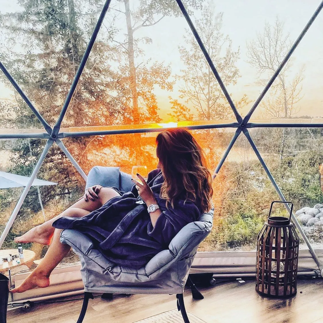 a woman in a bathrobe relaxing on a lounge chair inside the ignis dome enjoying a glass of white wine