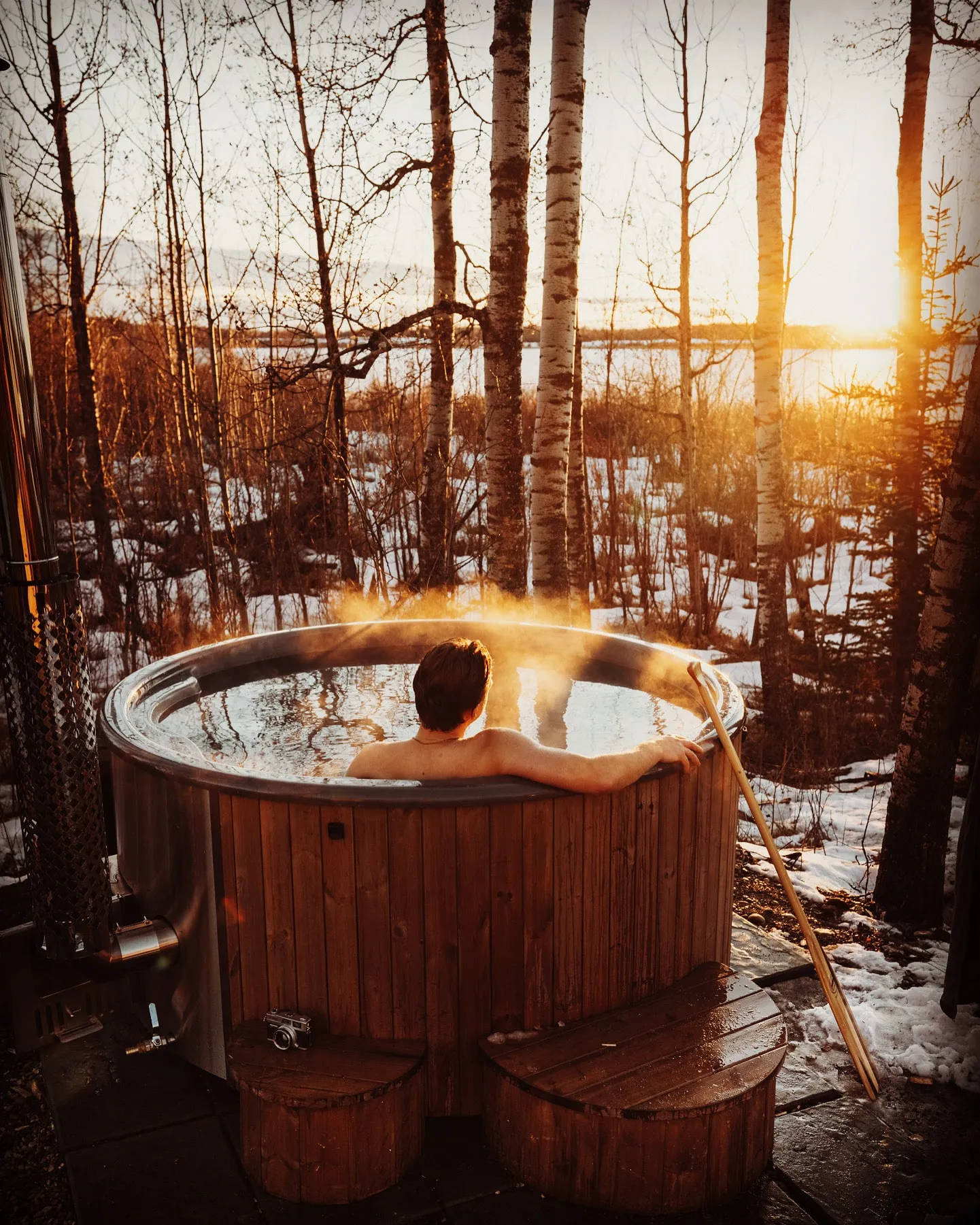 man sitting and relaxing in outdoor hot tub while looking forest and lake during a sunset.