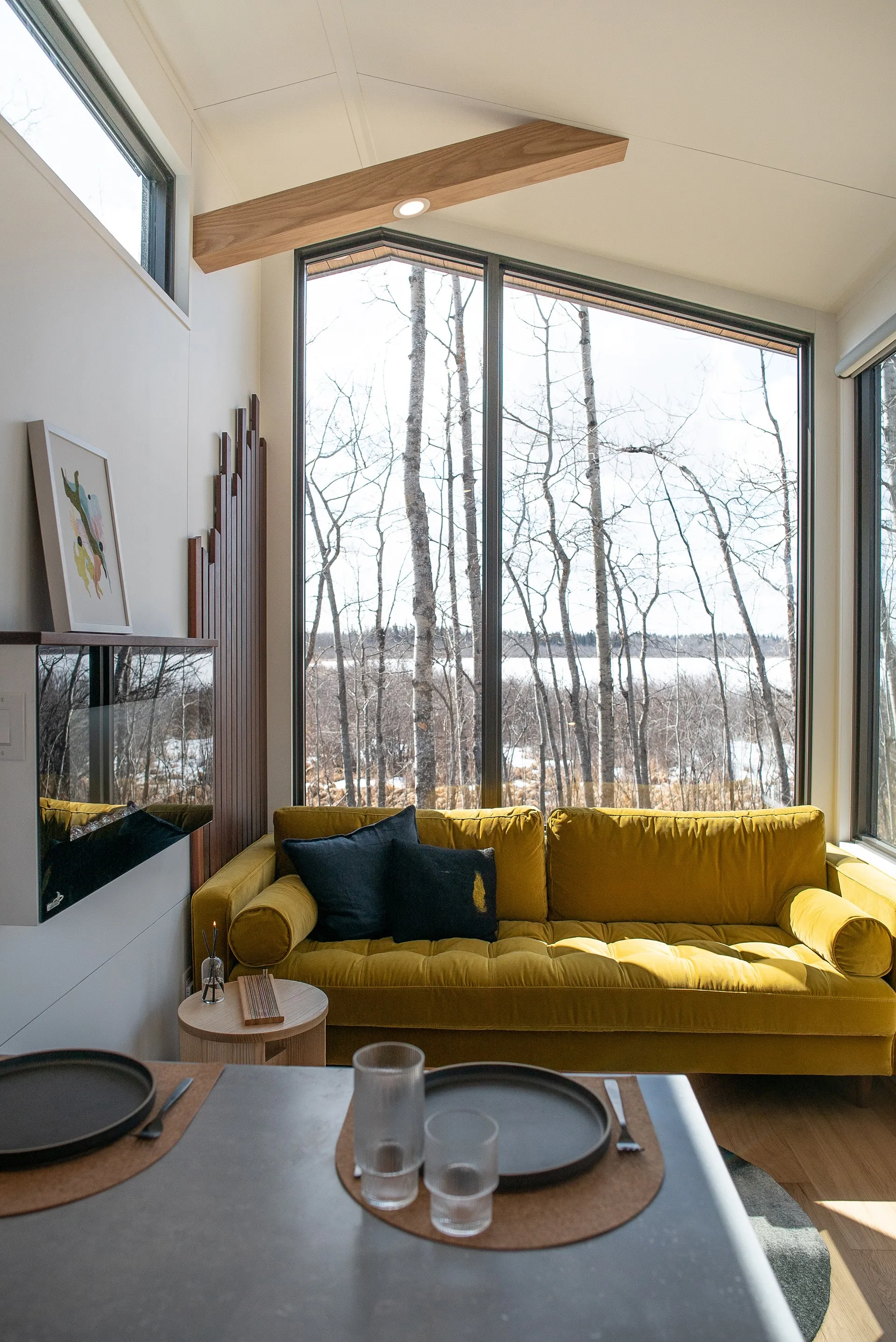 Kitchen counter view of Aqua tiny homes living room space with yellow double couch, floating fireplace, feature slat wall and large angled windows