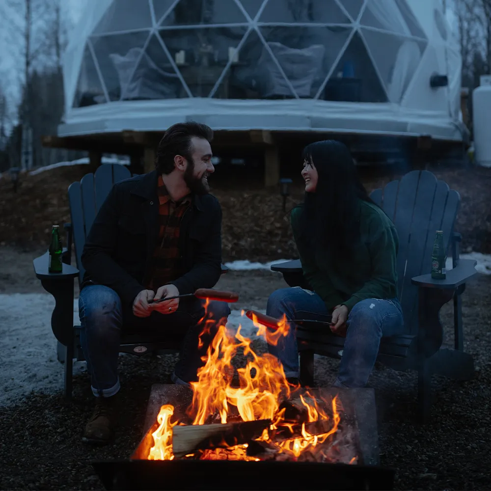 A couple enjoying each others company around the fire pit while sitting on patio chairs and roasting sausages and enjoying a beer with the ignis dome behind them